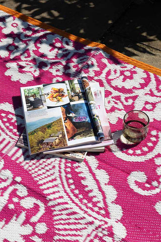 Close-up image of the Recycled Vintage Design Outdoor Rug in Pink styled with some magazines and a glass.