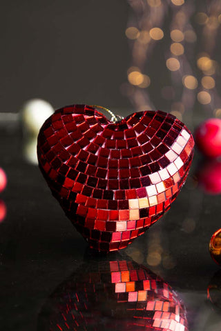 The Red Disco Heart Christmas Tree Decoration styled on a dark surface with baubles and fairy lights in the background.