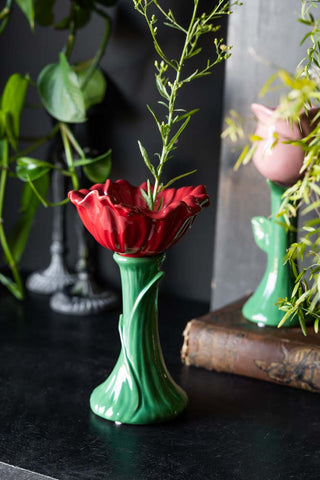 The Red Flower Earthenware Single Stem Vase styled with greenery inside, on a black surface with another vase and home accessories.