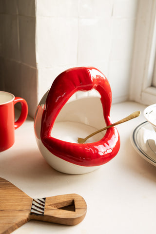 A lifestyle image of a red lips open mouth jar with sugar and a gold spoon in it. 