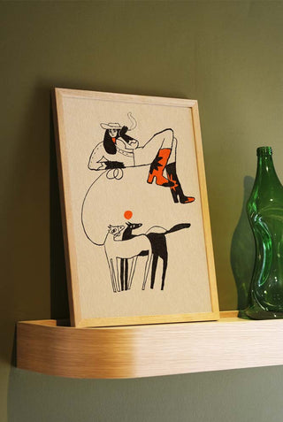 An unframed art print featuring a whimsical illustration of a woman in cowboy boots with red stars, sitting casually on a large abstract shape, with two stylized animals below. The print is displayed on a wooden shelf against a green wall with a green glass bottle beside it.