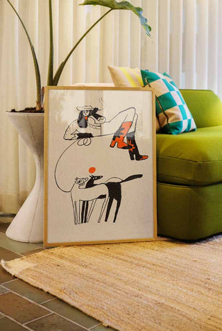 A stylish illustration print featuring a woman in striking red-starred boots, sitting atop a large abstract shape, with two stylized dogs below, displayed unframed on the floor next to a green sofa and a modern plant stand.