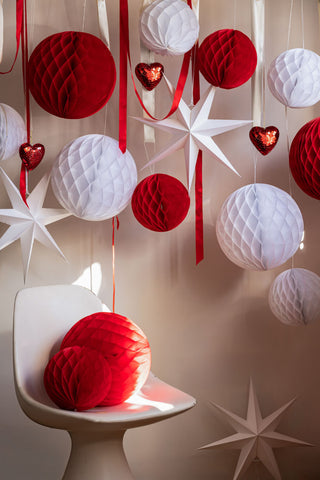 Lifestyle image of the Set Of 2 White Seven-Pointed Paper Star Decorations hung with red and white ribbon against a white wall.