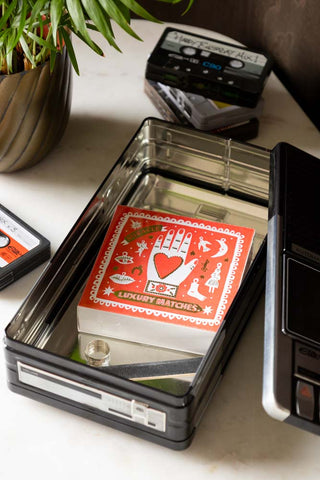 The Retro Cassette Recorder Storage Tin with the lid off, styled with some matches and a ring inside, displayed with a potted plant and some cassette tape storage tins.