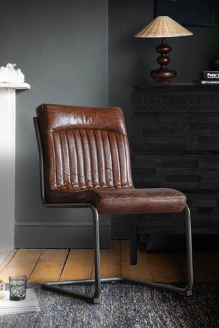 The Ribbed Antique-Style Brown Leather Chair displayed on a rug next to some magazines and a water glass, with a fireplace and a sideboard in the background.