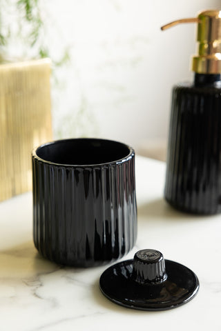 The Ribbed Black Glass Storage Canister with the lid off, displayed on a white marble surface with the matching soap dispenser in the background.