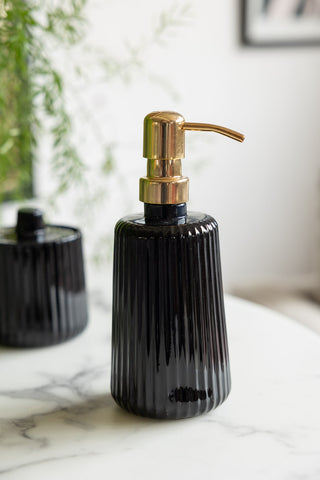 The Ribbed Black Glass & Gold Effect Soap Dispenser displayed on a white marble table with a matching storage pot and a plant in the background.