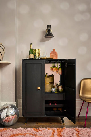 The Ringo Drinks Cabinet with one door open, styled with various bar and party accessories, next to a chair and a rug.