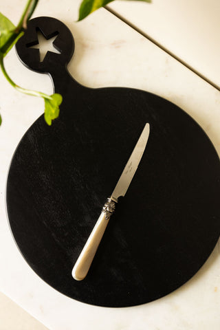 The reverse side of the Round Black Food Is My Love Language Serving Board displayed on a white table with a knife on and a plant in the shot.