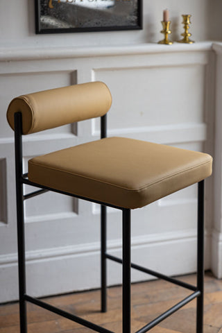 A cropped in image of the Sand Faux Leather Roll Back Bar Stool styled in front of a white panelled wall