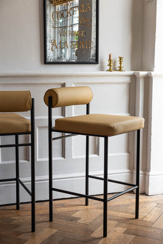 Two sand-coloured bar stools styled on wooden floor in front of a white wall, with a decorative mirror and some gold candlestick holders.