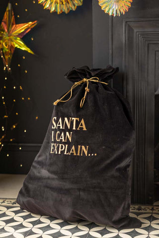 The Luxury Santa I Can Explain Velvet Christmas Sack styled in front of a black fireplace with decorations and fairy lights.