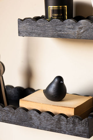 Close-up of the Set Of 2 Black Wooden Scalloped Wall Shelves displayed on the wall, styled with a candle, book and a bird ornament.