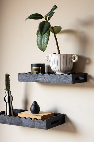 The Set Of 2 Black Wooden Scalloped Wall Shelves styled on a neutral wall, with various home accessories and a plant on.