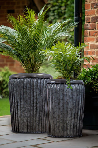 The Set Of 2 Galvanised Iron Ribbed Planters styled with plants inside on an outdoor patio.