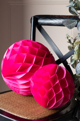 The Set Of 2 Hot Pink Honeycomb Ball Decorations styled on a black cross back chair next to a green garland plant.