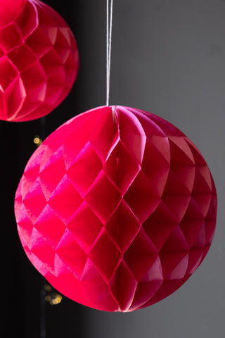 Close-up of one of the Set Of 2 Hot Pink Honeycomb Balls, styled hanging in front of a dark wall with fairy lights.