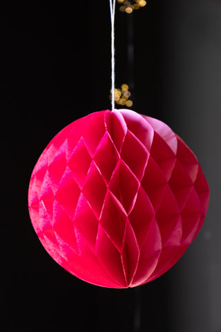 One of the Set Of 2 Hot Pink Honeycomb Balls hanging in front of a dark wall with fairy lights in the background.