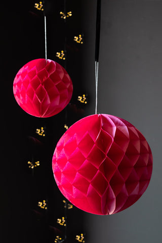 The Set Of 2 Hot Pink Honeycomb Balls hanging in front of a dark wall, styled with fairy lights.