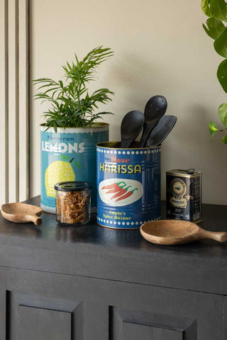 The Set Of 2 Lemon & Harissa Storage Tins - Large displayed together on a black sideboard, styled with plants, utensils and other kitchen accessories.