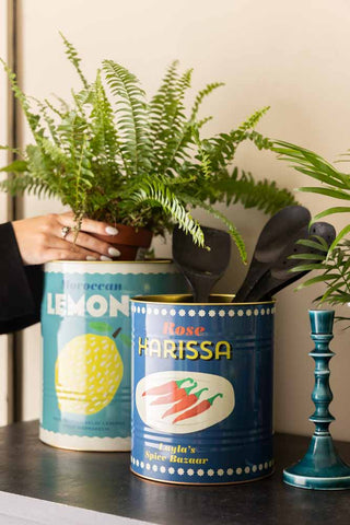 The Set Of 2 Lemon & Harissa Storage Tins - Large & Medium with a plant being places inside one, also styled with utensils and a candlestick.