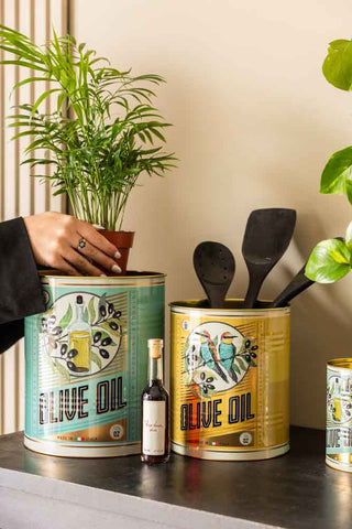 The Set Of 2 Olive Oil Storage Tins styled on a black sideboard, with a plant being put inside one, as well as utensils and other kitchen accessories around it.