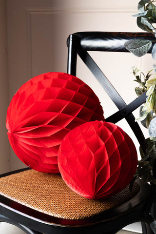 The Set Of 2 Red Honeycomb Ball Decorations styled on a black and rattan chair against a light pink backdrop.