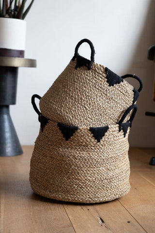 The smaller Seagrass Basket stacked on top of the large version, displayed on a wooden floor in front of a plant stand with plant and a desk chair.