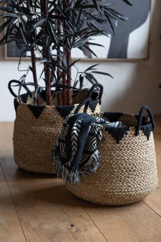 The Set Of 2 Seagrass Baskets styled on wooden floor in front of an art print on the wall, with a blanket in the smaller basket and a faux plant in the larger basket.