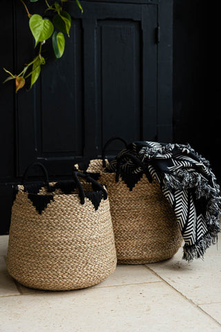 The Set Of 2 Seagrass Baskets styled on a tiled floor in front of a black sideboard with a blanket inside, with a branch of greenery in the background.