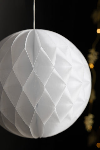 Close-up of one of the Set Of 2 White Honeycomb Balls, hanging in front of a black wall with fairy lights.