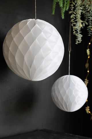 The Set Of 2 White Honeycomb Balls displayed in front of a black wall with greenery and fairy lights.