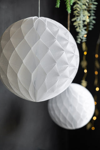 The Set Of 2 White Honeycomb Balls hanging in front of a black wall with greenery and fairy lights.