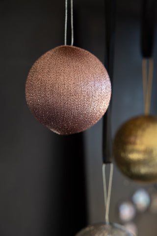 Close-up of the pink bauble in the Set Of 3 Metallic Spun Thread Christmas Bauble Decorations, hanging in front of a dark wall with the others in the background.