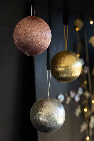 The Set Of 3 Metallic Spun Thread Christmas Bauble Decorations styled hanging in front of a dark wall, with floral decorations and fairy lights in the background.