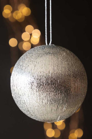 The Silver Metallic Spun Thread Christmas Bauble Decoration hanging in front of a dark wall with fairy lights.