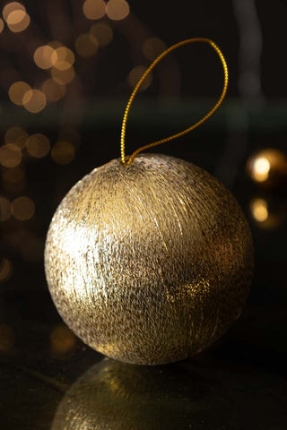 The Gold Metallic Spun Thread Christmas Bauble Decoration styled on a dark surface with fairy lights and decorations in the background.