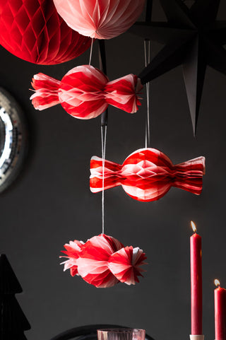 The Set Of 3 Pink & Red Honeycomb Sweets styled with other paper Christmas decorations in front of a black wall, with lit candles.