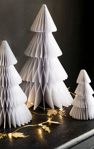 A lifestyle image of the Set Of 3 White Paper Trees, displayed on a black surface with some fairy lights in the background.