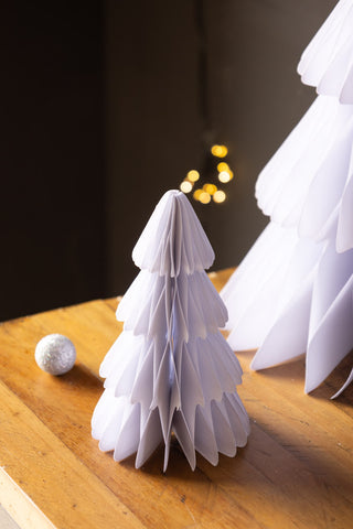 Two of the Set Of 3 White Paper Trees, displayed on a wooden surface with a bauble and some fairy lights in the background.