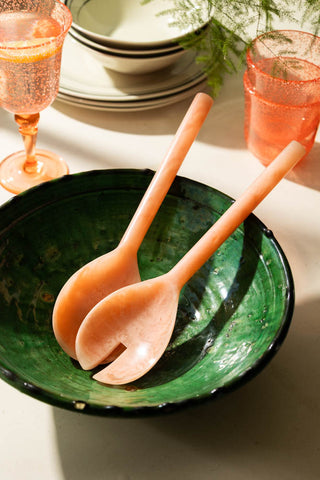 A lifestyle image of pink salad servers in an emerald green ceramic bowl. 