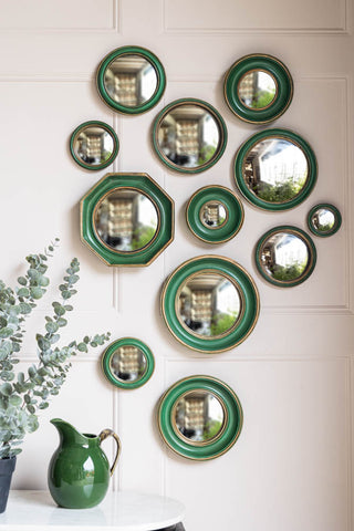 The Set of 12 Antique Green & Gold Convex Framed Mirrors styled on a light wall, above a table with a green jug and a plant on.