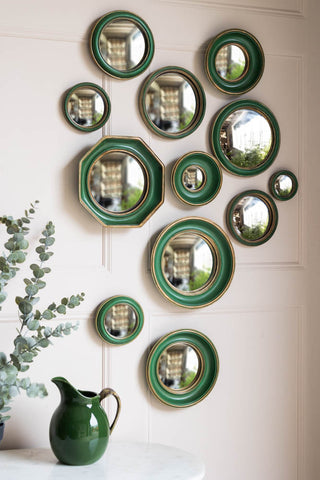 The Set of 12 Antique Green & Gold Convex Framed Mirrors displayed on a white panelled wall, above a table styled with a green jug and a plant on.