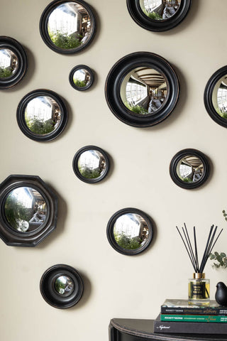 Close-up of the Set of 12 Black Convex Framed Mirrors styled on a wall, above a table with some books, a diffuser, a bird ornament and a plant.