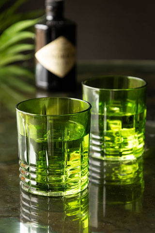 The Set of 2 Green Whiskey Embossed Glasses styled on a counter, with a plant and a bottle in the background.