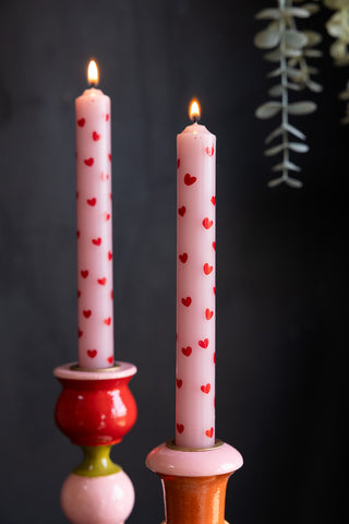 The Set of 2 Red & Pink Heart Dinner Candles lit and styled in candlestick holders, in front of a dark wall and plant.