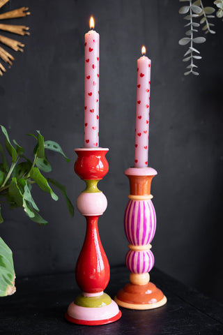 The Set of 2 Red & Pink Heart Dinner Candles lit and styled in colourful candlestick holders, styled on a sideboard in front of a dark wall with plants and an ornament.