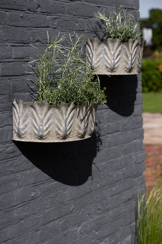 The Set of 2 Rustic Wall Planters displayed outside on a dark brick wall with plants inside.