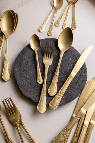 An antique-style gold cutlery set, styled against a marbled dark grey plate on a white tablecloth 