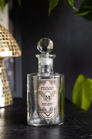 One of the Set of 3 Apothecary-Style Poison Decanter Bottles, styled on a black surface with an ornament and a plant in the background.